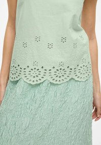 Mentolově zelený top s květinovým výřezem na krajkovém lemu, nad texturovanou mentolově zelenou sukní s krčenou látkou. Oba prvky jsou měkké a vrstvené.