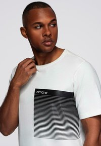 Bílý krátký rukáv tričko s černým obdélníkovým grafickým prvkem s horizontálními pruhy a slovem "ombre" vytištěným nahoře.