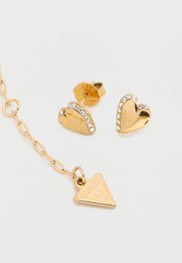Σετ κοσμημάτων σε χρυσό τόνο με σκουλαρίκια σε σχήμα καρδιάς διακοσμημένα με στρας, και τριγωνικό μενταγιόν με λογότυπο σε αλυσίδα.