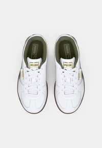 Zapatillas de cuero blancas con perfil bajo, punta redonda, forro interior verde y detalles dorados en la lengüeta y el costado. Suela de goma.