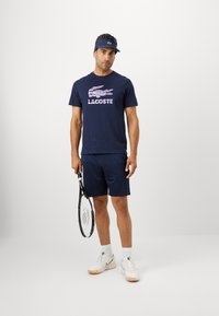 Άνδρας σε σπορ νavy ένδυση και καπέλο, κρατώντας μια ρακέτα τένις, σε φόντο λευκό.