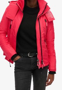 Jasnoróżowa pikowana kurtka z wysokim kołnierzem, czarnymi akcentami i wieloma kieszeniami na suwak. Pod spodem teksturowany czarny sweter. Czarny pasek.