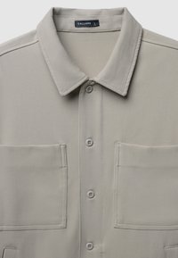 Camicia beige chiaro a maniche corte con colletto, due tasche frontali e cinque bottoni. Tessuto liscio con una sottile texture e design su misura.