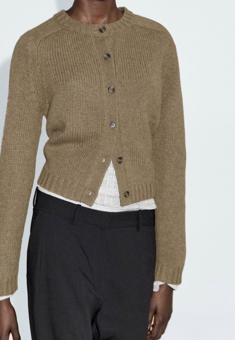 Personne portant un cardigan court marron tricoté avec des boutons, une chemise blanche texturée en dessous et un pantalon noir taille haute.