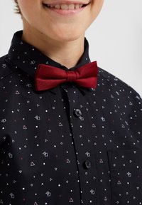 Camisa negra con patrones geométricos en blanco y rojo, que presenta una pajarita de satén roja y un cuello abotonado. Bolsillo en el lado izquierdo.