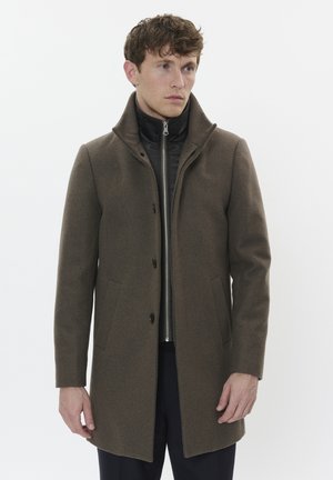 Mann in einem braunen Wollmantel, der über einer schwarzen Zip-Jacke getragen wird, steht vor einem schlichten weißen Hintergrund.