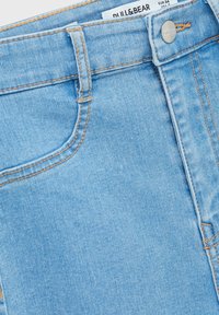 Vaqueros de denim azul claro con diseño de cinco bolsillos, costuras naranjas y un botón metálico de marca en la cinturilla.