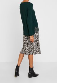 Dunkelgrünes Langarmoberteil mit einer Spitzenakzent an der Seite, kombiniert mit einem beigefarbenen Midi-Rock im Leopardenmuster und schwarzen Stiefeletten.