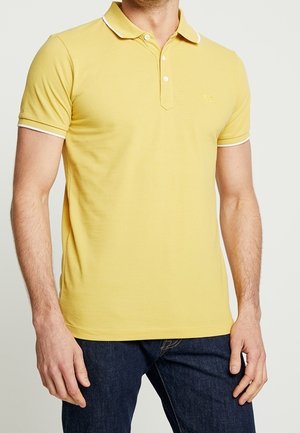 Homme portant un polo jaune cintré à manches courtes avec bordure blanche au col et aux manches, associé à un jean bleu foncé.