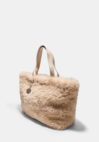 Tote taška z umelého kožušiny v svetlej béžovej farbe, s jemným, textúrovaným povrchom, béžovými koženými rúčkami a strieborným akcentom v podobe tagu.