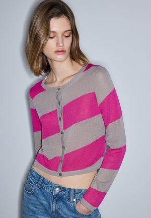 Vrouw met schouderlang haar kijkt omlaag, draagt een cropped vest met brede diagonale strepen in roze en grijs en een blauwe spijkerbroek, met één hand in de zak.