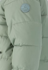 Ljusgrön pufferjacka med kviltat mönster, med en rund logopatch och slät yta, visad från en närbildsvinkel.