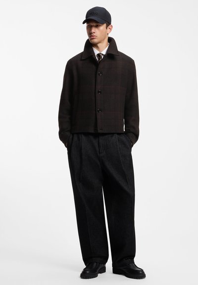 Jeune homme portant une veste noire boutonnée, un pantalon large foncé, des chaussures noires, une cravate, une chemise blanche et une casquette noire, debout les mains dans les poches.