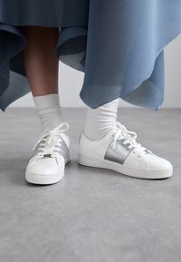 Sneakers bianchi con dettagli e lacci argentati, caratterizzati da una finitura in pelle liscia. Presentano un design semplice, low-top e una punta arrotondata.
