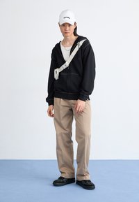 Felpa nera con zip, canottiera bianca, pantaloni beige, scarpe nere. Cappellino bianco con la scritta "DICKIES." Borsa con fascia regolabile sulla parte anteriore del corpo.