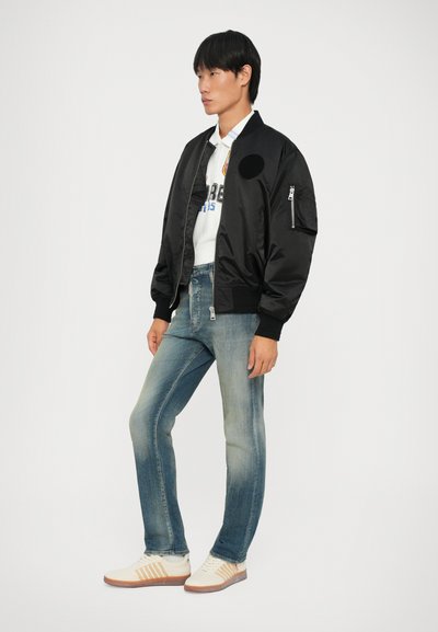 Giubbotto bomber nero con finitura satinata, polsini a costine, dotato di una tasca sulla manica sinistra e abbinato a jeans in denim blu chiaro e sneakers beige.