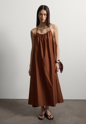 Donna con capelli lunghi e lisci che indossa un vestito marrone senza maniche lungo, sandali neri e una sciarpa bordeaux legata intorno al polso, in piedi su un pavimento in cemento.