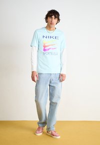 Šviesiai mėlynas Nike marškinėlis su spalvingu logotipu, uždėtas ant baltų ilgų rankovių marškinių, derinamas su šviesiai mėlynomis džinsais ir rožiniais sportbačiais.