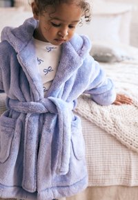 Peignoir doux violet clair avec capuche, ceinture à nouer et deux poches avant. Porté sur un haut blanc avec des motifs de nœuds bleus. Texture douce.