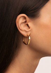 Primer plano de la oreja de una mujer con el cabello lacio castaño, que lleva un arete de aro dorado retorcido y brillante, contra un fondo neutro.