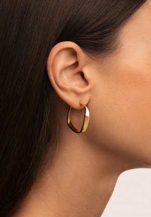 Primo piano di un orecchio femminile con capelli castani lisci che indossa un orecchino a cerchio dorato lucido e attorcigliato su uno sfondo neutro.