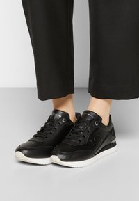 Czarne skórzane sneakersy niskie z białymi podeszwami i czarnymi sznurówkami, noszone z czarnymi spodniami 3/4 na szarym podłożu.