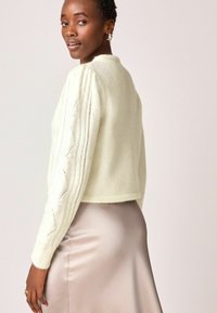 Pulover din tricot ivory cu mâneci texturate, cu model în cablu; design scurt. Asortat cu o fustă din mătase, de un bej deschis.