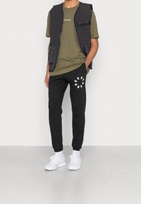 Gilet imbottito nero con tasche laterali sopra una maglietta verde oliva. I pantaloni della tuta neri presentano una stampa di uccelli bianchi. Le sneaker bianche completano l'outfit.