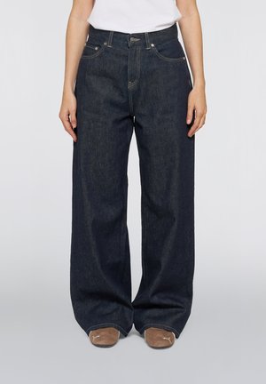 Person iført mørkeblå vidde jeans og brune sko, stående med hænder afslappet ned langs siderne mod en ensfarvet baggrund.