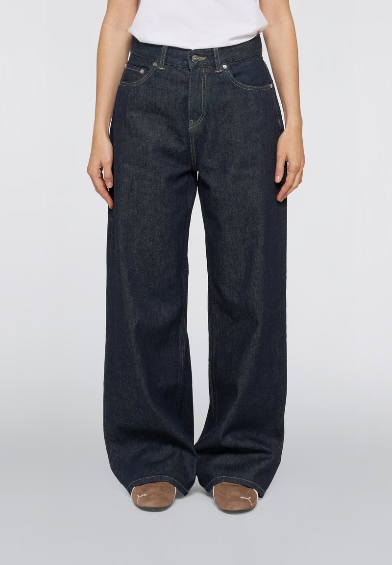 Personne portant un jean large bleu foncé et des chaussures marron, debout, les mains détendues le long du corps, sur un fond uni.