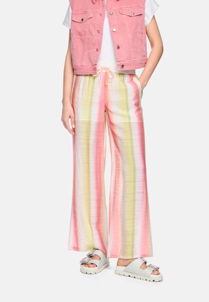 Personne portant un gilet en denim rose pastel, un haut blanc, un pantalon large à rayures verticales jaunes et roses, et des sandales blanches avec des nœuds.