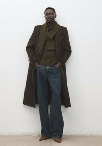 Brązowy wełniany płaszcz na zielonym dzianinowym swetrze, zestawiony z ciemnymi rozszerzanymi dżinsami i brązowymi szpiczastymi szpilkami. Gładkie tekstury i neutralna paleta kolorów.