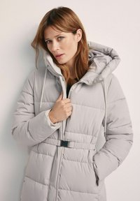 Jasnoszary puchowy płaszcz z odpinanym kapturem, zapięciem na zamek, sznurkiem ściągającym i paskiem w talii. Posiada poziome pikowanie i boczne kieszenie.