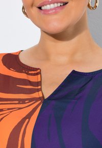 Blusa colorata con scollatura a V, caratterizzata da audaci motivi arancioni, marroni e viola. Tessuto morbido e dettagli di cucitura visibili.