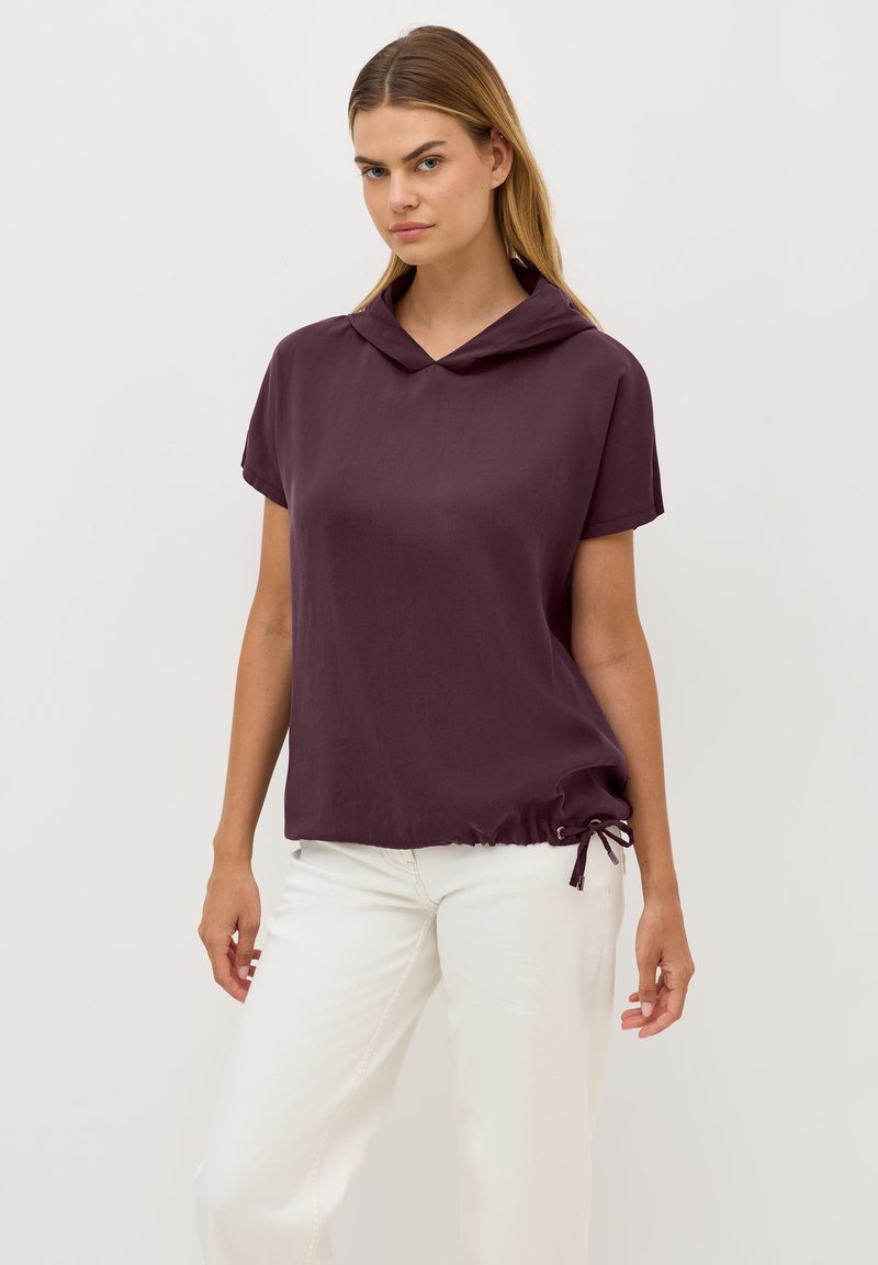 Haut à manches courtes et à capuche de couleur violet profond avec un ourlet à cordon. Tissu lisse, coupe décontractée, associé à un pantalon blanc pour le contraste.