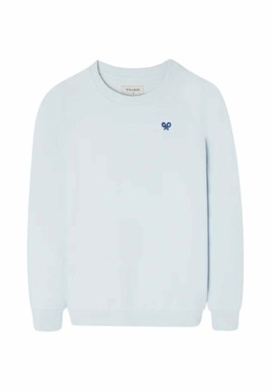 Niño que lleva un suéter azul claro sobre una camisa vaquera, pantalones cargo beige, zapatillas azul marino y una gorra de béisbol blanca y azul, de pie mirando hacia adelante.