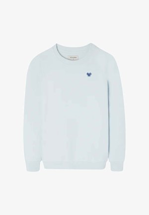 Niño que lleva un suéter azul claro sobre una camisa vaquera, pantalones cargo beige, zapatillas azul marino y una gorra de béisbol blanca y azul, de pie mirando hacia adelante.