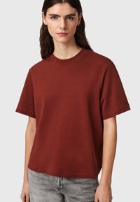Femme aux longs cheveux bruns portant un t-shirt uni à manches courtes de couleur rouille et un jean gris clair sur un fond neutre.