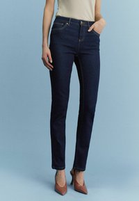 Tummansininen farkkuhousu slim fit -mallilla, joissa on vetoketjukiinnitys, viisi taskua ja kontrastoiva oranssi ommel. Yhdistetty ruskeiden korkokenkien kanssa.