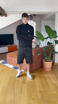Noor inimene lühikeste juustega, mustas lukuga tuulejope, mustades lühikeses ja valgetes sneakers'ites seisab risti käed moodsa elutoas.