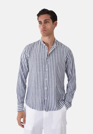 Uomo che indossa una camicia a righe verticali grigie e bianche con bottoni, colletto alla coreana e pantaloni bianchi, in piedi con una mano in tasca.