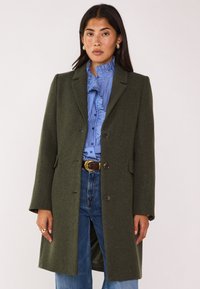 Cappotto di lana verde oliva con un design su misura, rever a scollatura, e tre bottoni. Presenta tasche laterali e un tessuto leggermente strutturato.