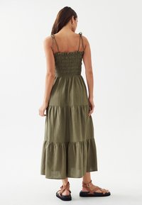 Olivenfarbenes Maxikleid mit gerafftem Oberteil, verstellbaren Trägern und gestuftem Rockdesign. Kombiniert mit braunen Riemchensandalen.