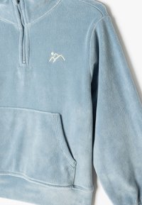 Fleece pullover in lichtblauw met een ritskraag tot de helft, kangoeroezak en geborduurd berglogo in het wit. Zachte textuur.