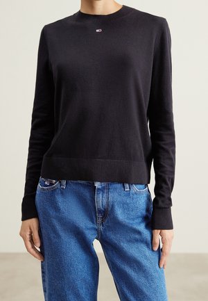 Person iført en tætsiddende sort rundhalset sweater og blå denimjeans, stående med hænderne afslappet ned langs siderne.