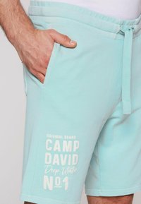 Lichtblauwe shorts van zachte stof, met zijzakken en een trekkoord in de taille. Witte tekstgrafieken op het linkervoorbeen.