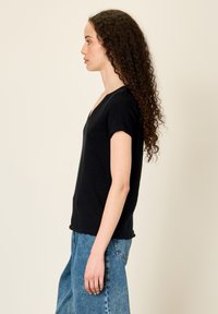 Žena s dlouhými kudrnatými vlasy, oblečená v černém krátkém tričku s véčkovým výstřihem a modrých džínách, stojící bokem proti jednoduchému pozadí.