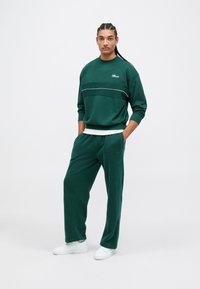 Homem com cabelo trançado vestido com fato de treino verde escuro e ténis brancos, de pé com as mãos nos bolsos contra um fundo branco liso.