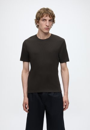 Ung mand med krøllet blondt hår iført en ensfarvet sort t-shirt med korte ærmer og mørke bukser, stående mod en hvid baggrund.