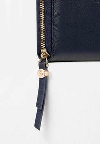 Marinblå läderclutch med texturerad yta, guldzipper och hårdvarudetaljer. Har en rem med en varumärkespräglad dekorativ detalj.